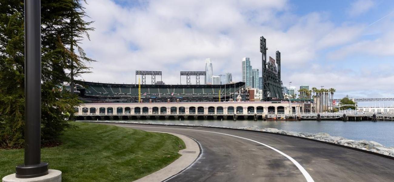 Paved pathway leads around China Basin Park with clear views of Oracle Park across the waterway.