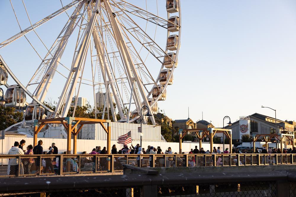 A crowd of people standing on the Little Embarcadero at Fisherman's Wharf near the Skystar Wheel.
