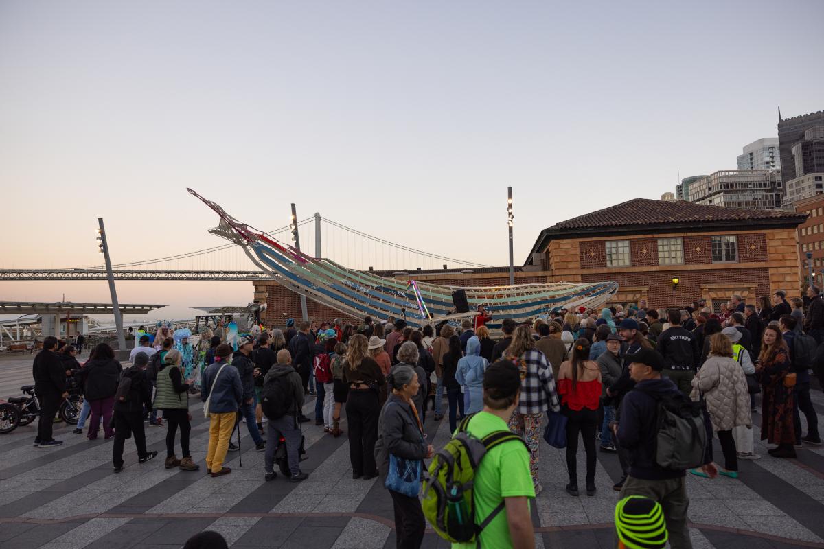 A crowd stands around a sculpture of a large blue whale installed at the Downtown Ferry Terminal Plaza.