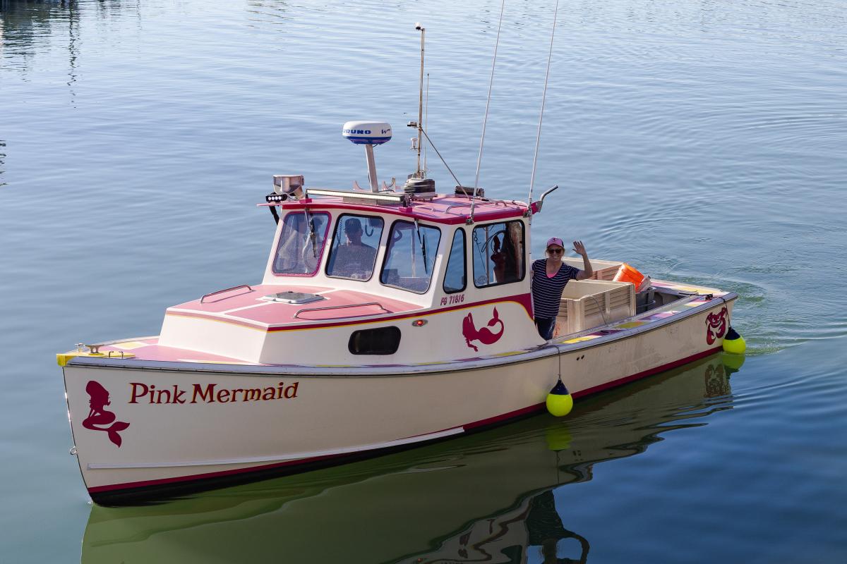 Captain Holly Fruehling waves from aboard her fishing boat, Pink Mermaid. A man is visible in the boat cabin.