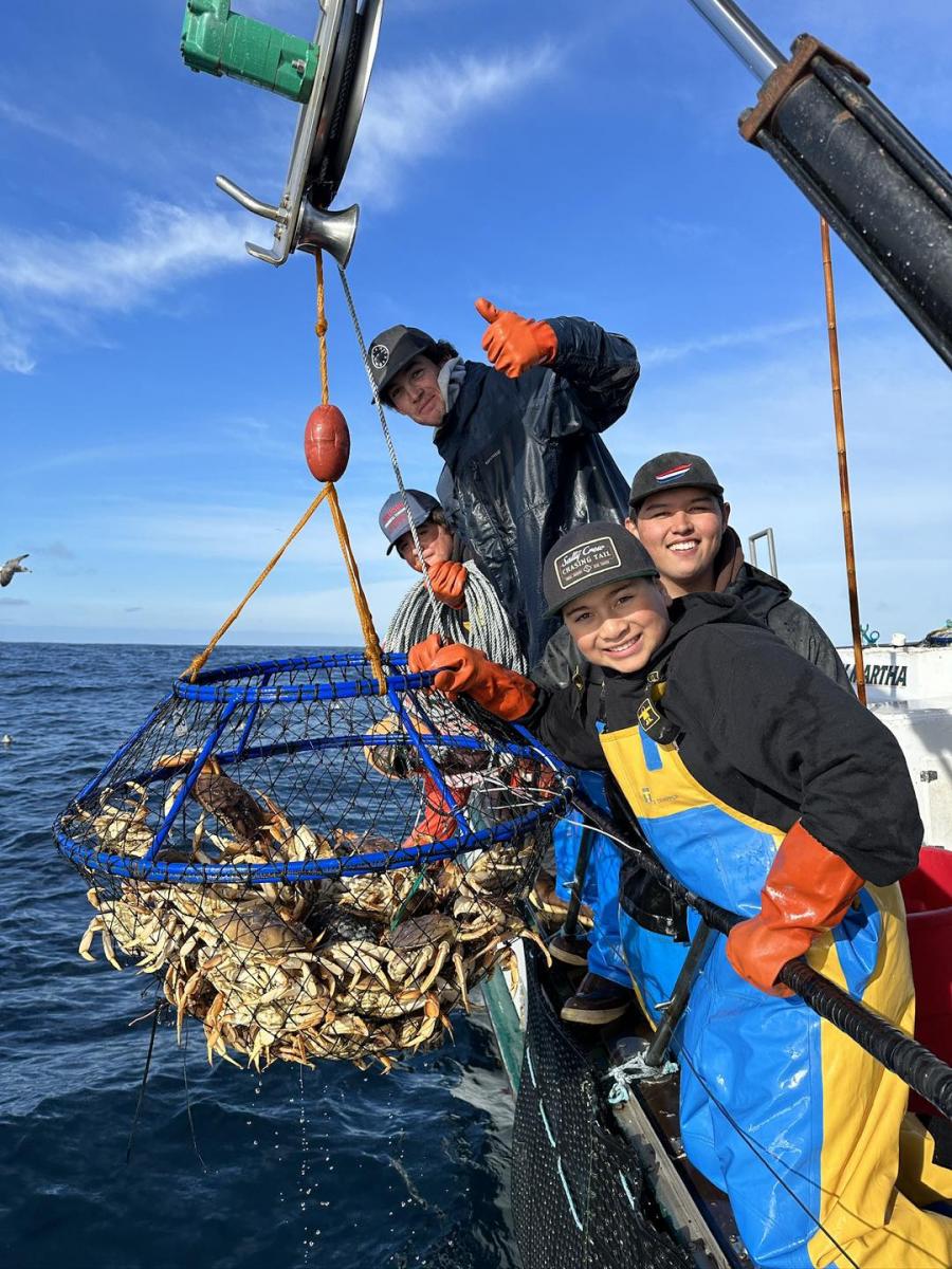 Group of children and adults smile aboard a boat, pulling up a crab pot.