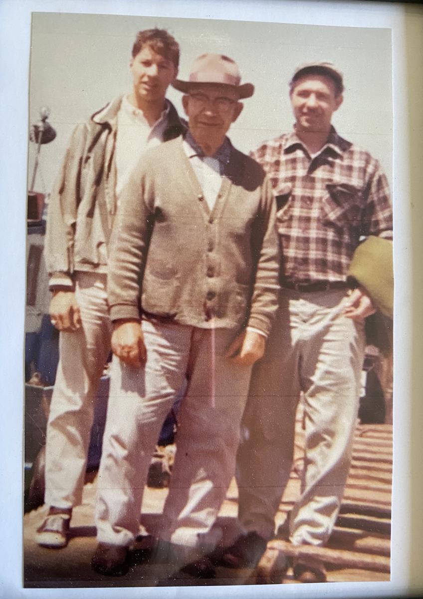 Three men of different ages standing in front of a boat. 