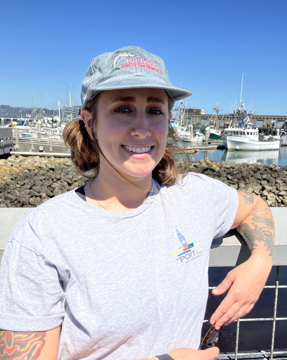 Becca Gross smiles in front of Hyde Street Harbor, wearing a Port of San Francisco tee shirt. 