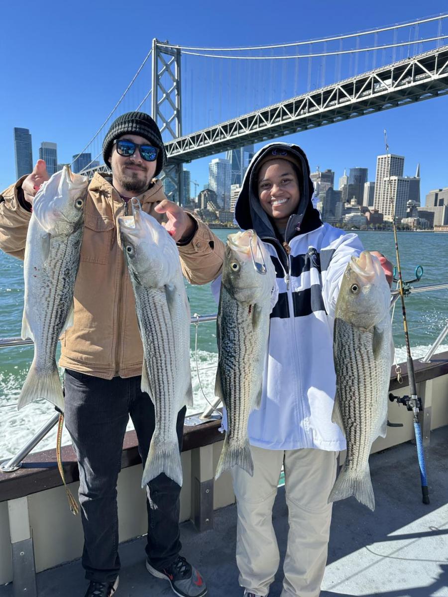 Two young people hold up large striped bass on a boat in front of the Bay Bridge. 