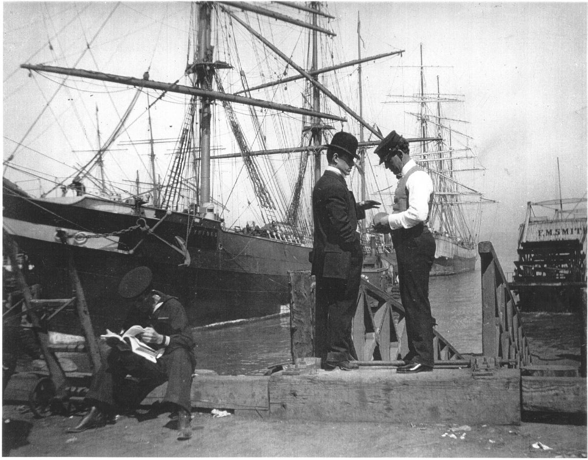 Two men in vintage clothing stand on a pier negotiating with boats in the background. 